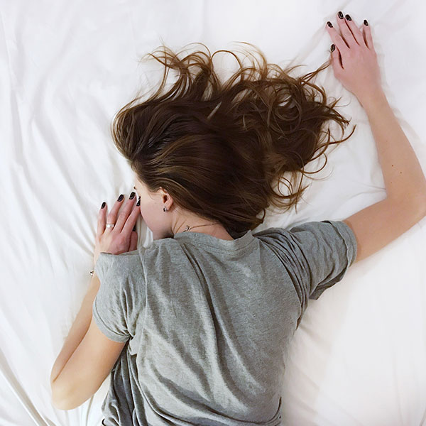 A woman lies on her stomach on a bed with white sheets, wearing a grey t-shirt.