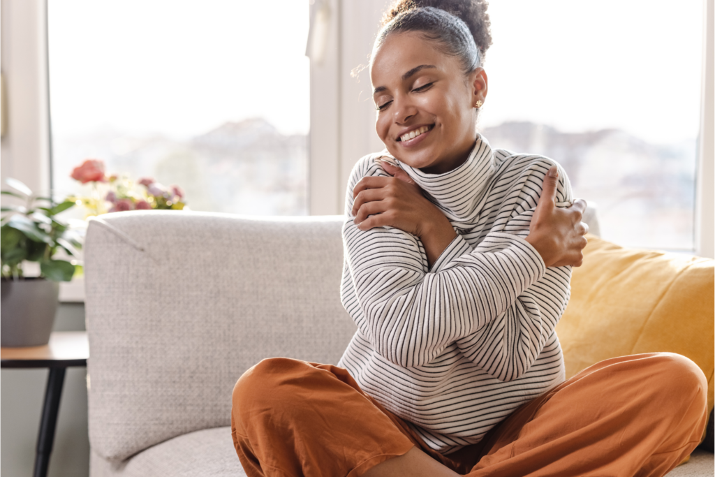 A woman sits on her white couch in her living room with large bay windows at her back hugging her arms around herself.